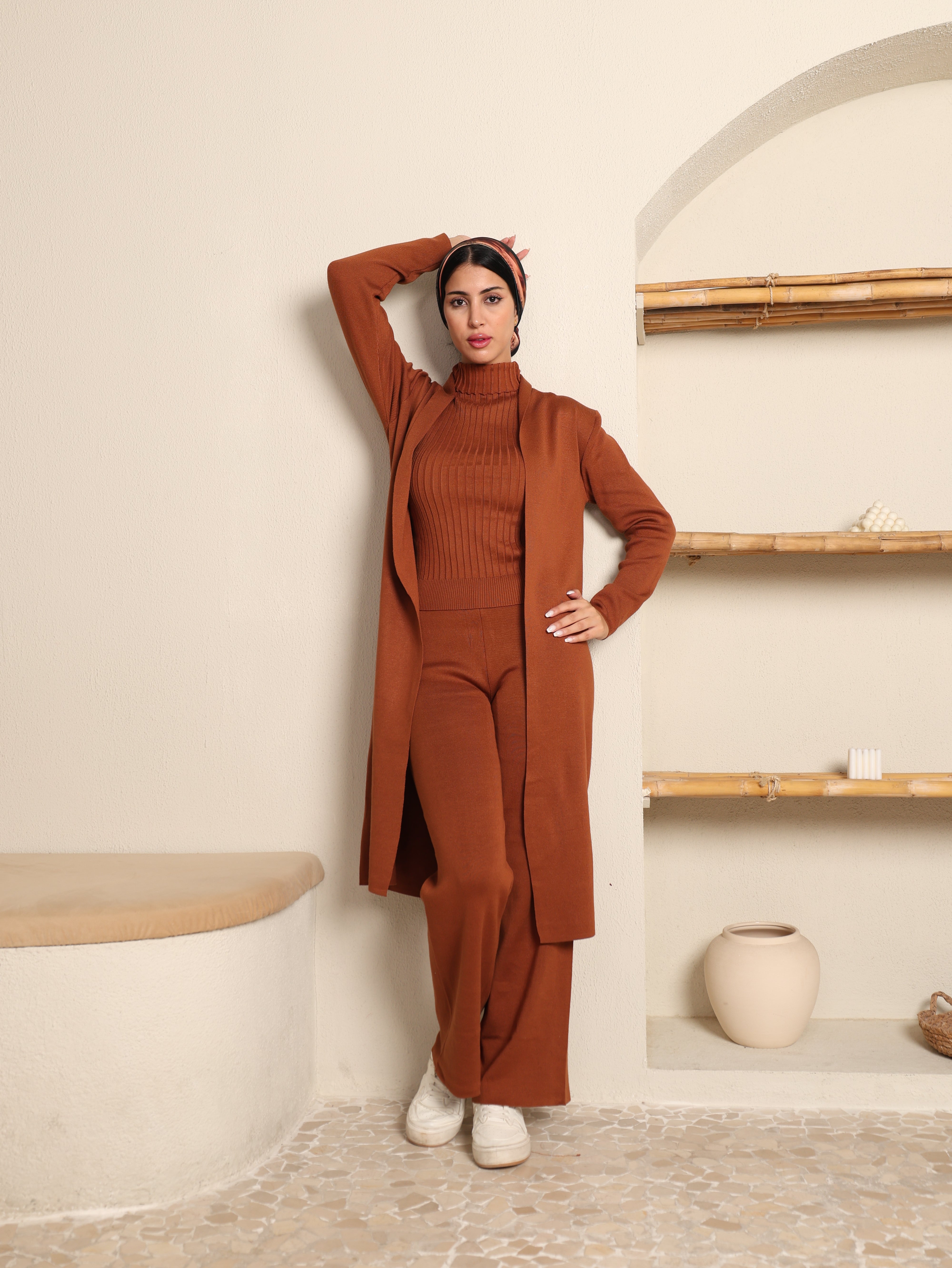 Person wearing a brown outfit standing in a minimalistic room with white walls and light-colored floor.